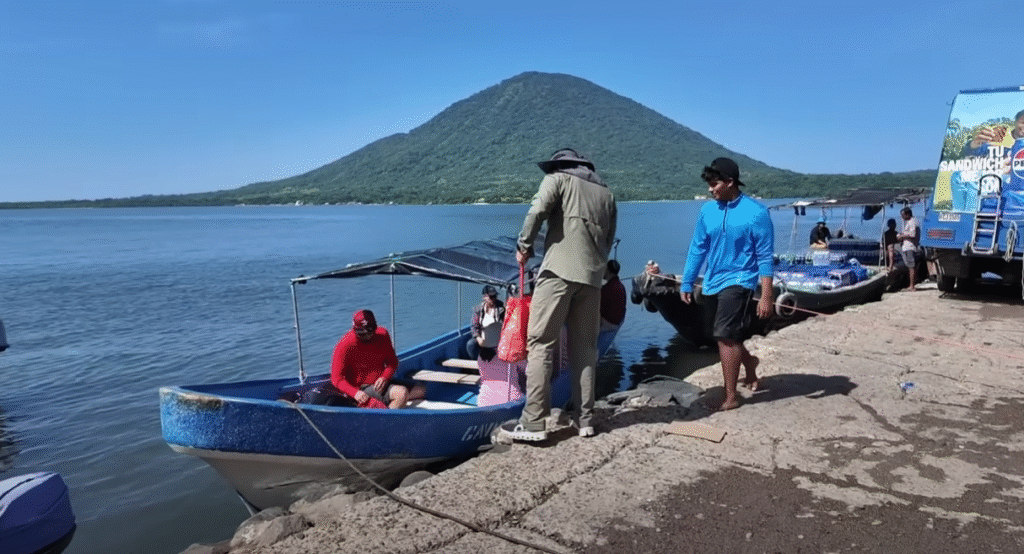 Personas abordando lanchas en un muelle del Golfo de Fonseca, con un volcán verdoso al fondo.