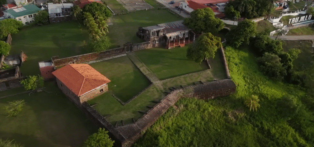 Vista aérea del Fuerte de Santa Bárbara en Trujillo, una fortificación histórica de muros de piedra que rodean una pequeña casa de techo rojo y un amplio jardín verde, ubicada junto a la costa.