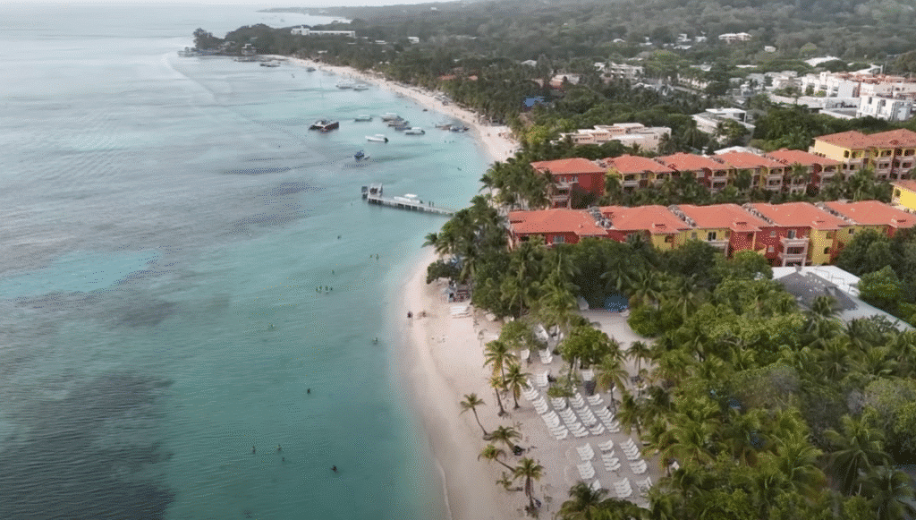 Vista aérea de un resort frente a la playa en Roatán con edificios de techo rojo, palmeras, arena blanca y aguas turquesas donde nadan turistas.