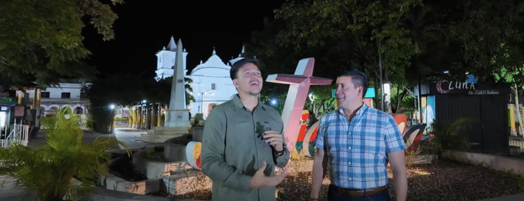 Dos hombres conversando en una plaza de Olancho por la noche, con una iglesia de fachada blanca y una cruz roja decorativa al fondo.