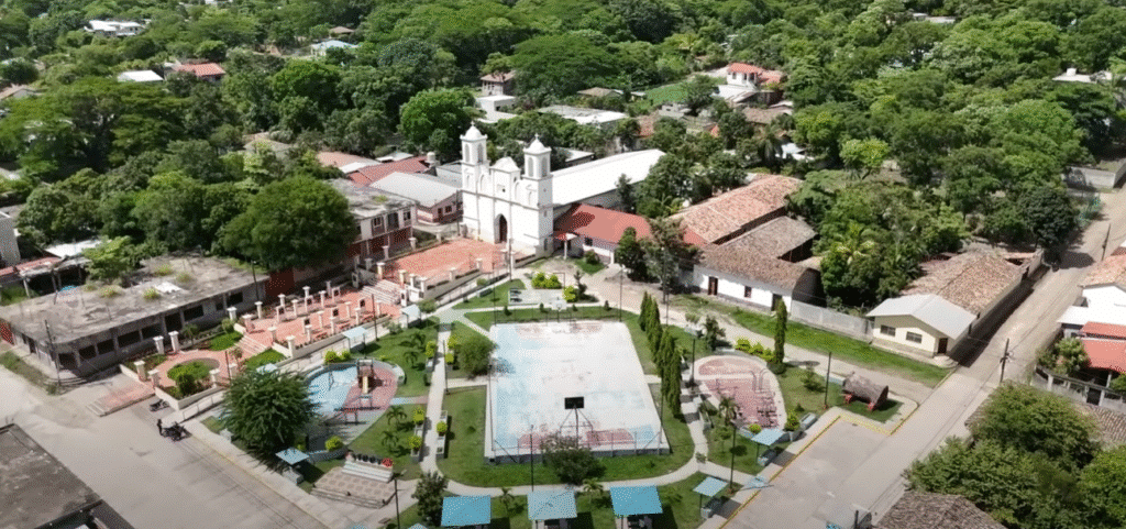Vista aérea de la plaza central de Intibucá con una iglesia colonial blanca de dos torres, una piscina o pila en la plaza y casas de techo rojo rodeadas de árboles.
