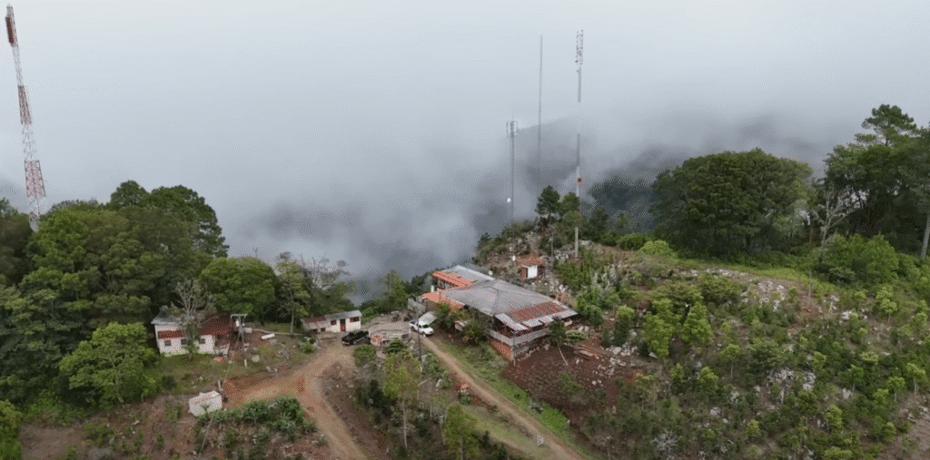 Vista aérea de una estación de antenas en la cima de una montaña en Comayagua, con torres de comunicación, edificios con techo rojo y caminos de tierra entre la vegetación, rodeada de nubes y niebla.