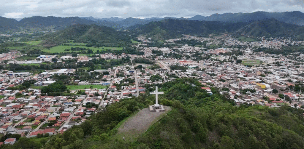 Vista aérea de Danlí con un gran mirador de cruz blanca en una colina que domina la ciudad; casas, calles y montañas se extienden al fondo.
