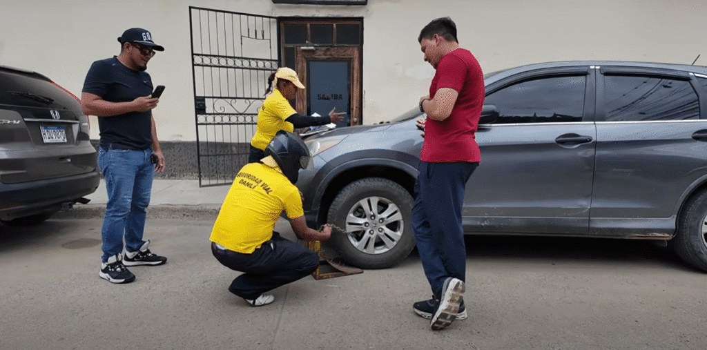 Grupo de jóvenes en Danlí arreglando un neumático de coche. Un hombre con camiseta amarilla y gorra negra está agachado revisando la llanta mientras otro con camiseta roja observa. Un tercer hombre con gorra azul revisa su teléfono y una mujer con sombrero amarillo está detrás del automóvil.