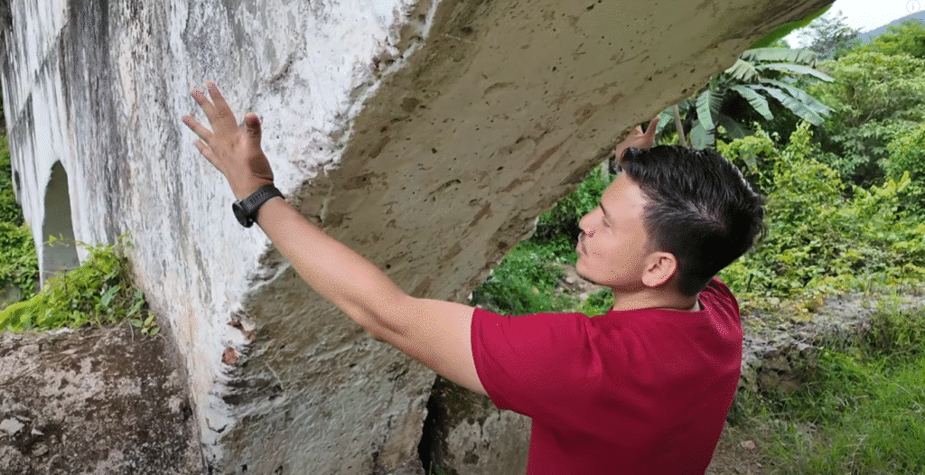 Hombre con camiseta roja explorando una ruina de arco de piedra en Danlí, apoyando sus brazos sobre la estructura con vegetación al fondo.