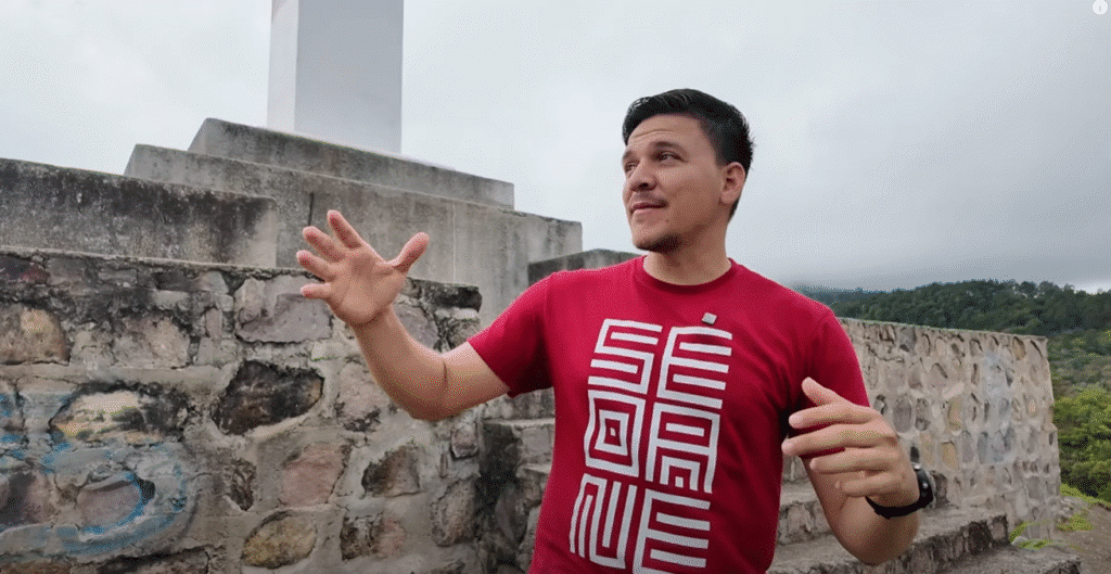Hombre con camiseta roja y diseño geométrico gesticulando frente a una pared de piedra en Danlí; al fondo, colinas verdes y cielo nublado.