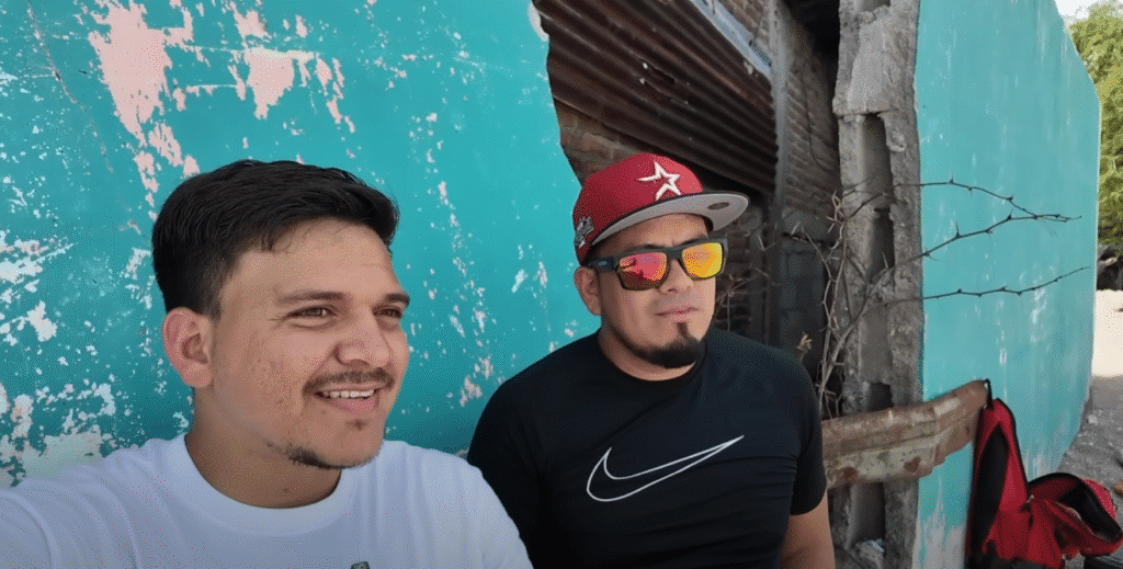 Dos hombres jovenes posan frente a una pared turquesa desgastada; uno lleva camiseta blanca y sonrie, el otro viste camiseta negra con gorra roja y gafas de sol.