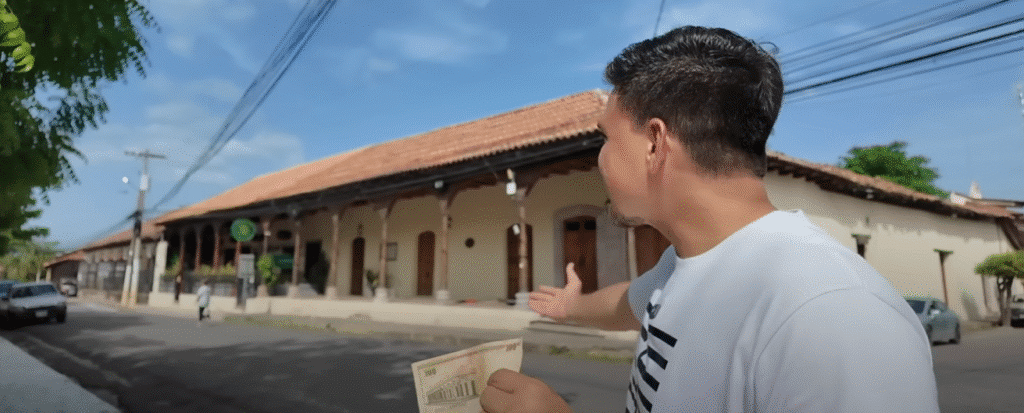 Hombre de perfil con camiseta blanca sosteniendo billetes de dinero frente a una antigua estacion o edificio colonial en Choluteca, con cielo azul y arboles.
