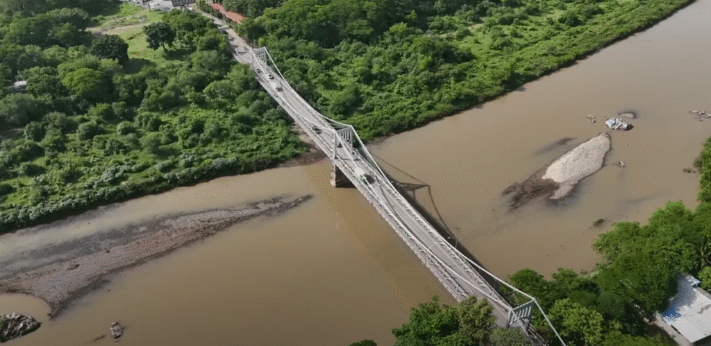 Vista aerea de un puente de metal cruzando un rio caudaloso de aguas marrones rodeado de vegetacion verde.
