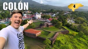 Selfie de un hombre sonriendo frente al Fuerte de Santa Bárbara y la iglesia de Trujillo, con la ciudad de Colón y montanas al fondo; el número 4 identifica la región.